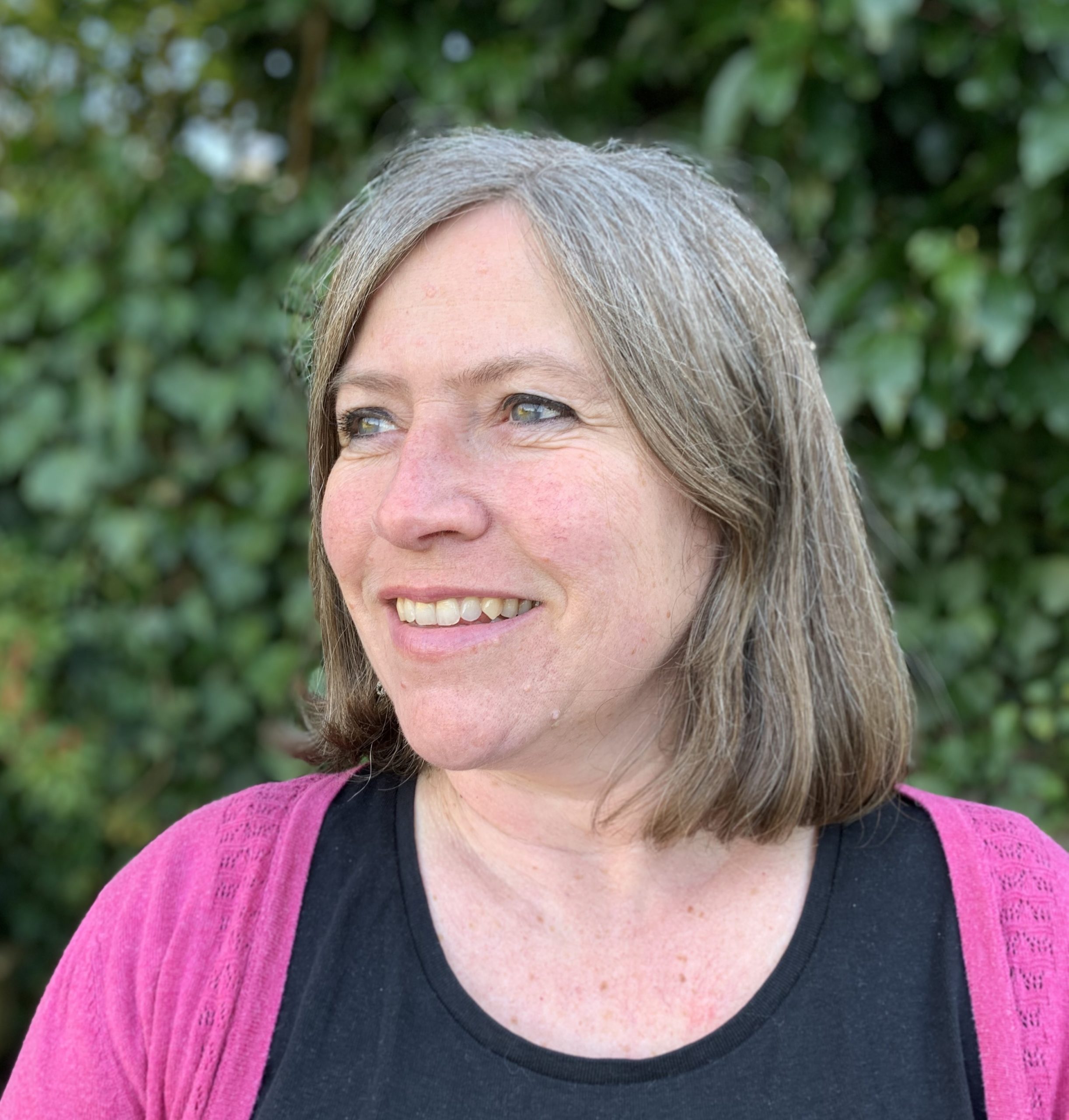 Image of Jane, editor. She is a white woman standing outside with a hedge behind her
